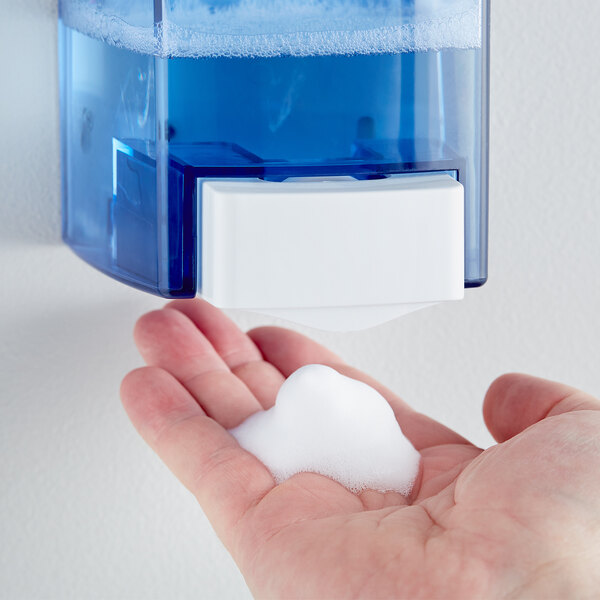 A hand holding a blue Noble Chemical foaming hand soap dispenser with white soapy foam.