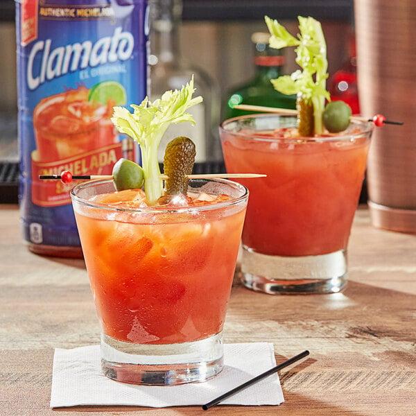 Two glasses of Clamato Michelada cocktails with pickle garnishes on a table.