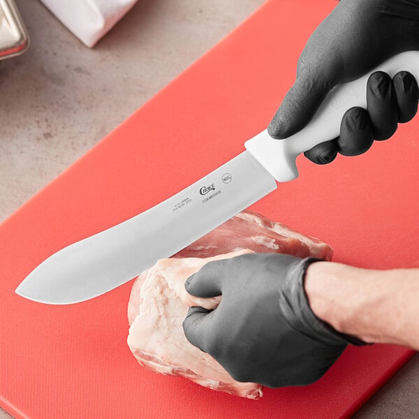 A person wearing a black glove using a Choice 8" Butcher Knife with a white handle to cut meat on a cutting board.