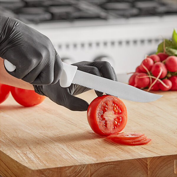 A person in gloves cutting a tomato in half with a Choice 8" serrated edge utility knife.