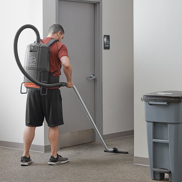 A man using a Hoover HVRPWR cordless backpack vacuum cleaner to vacuum the floor of a room.