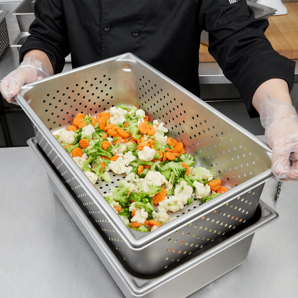 A person in a chef's uniform holding a Vollrath stainless steel pan full of vegetables.