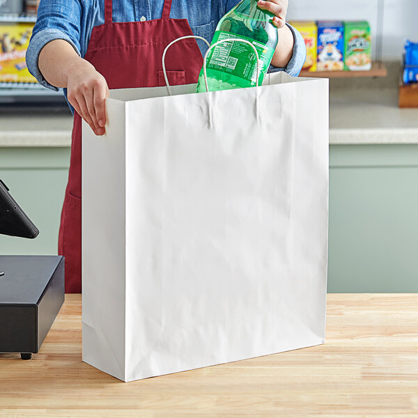 A person putting a green bottle into a white paper shopping bag with handles.
