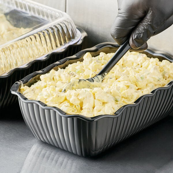 A gloved hand uses a spoon to serve potato salad from a black deli crock.