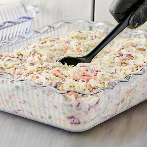 A person using a black spoon to serve coleslaw from a clear deli crock.