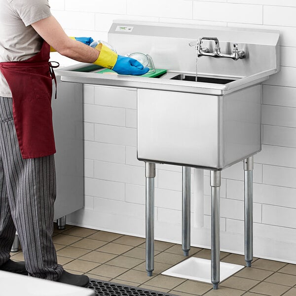 A man in a white shirt and blue gloves washing dishes in a Regency stainless steel sink with right drainboard.