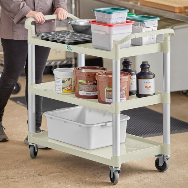 A woman pushing a beige Choice utility cart with food containers on it.