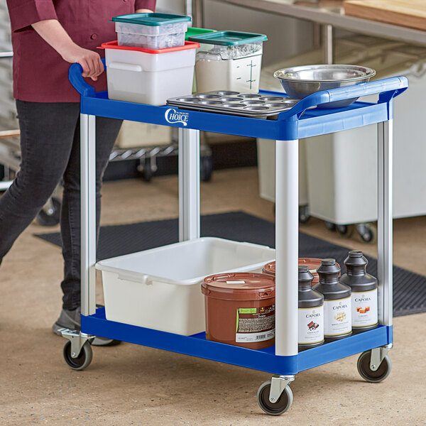 A woman pushing a blue Choice utility cart with containers on it.
