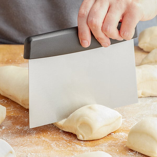 A person using a Choice stainless steel dough cutter with a black handle to cut dough on a table.