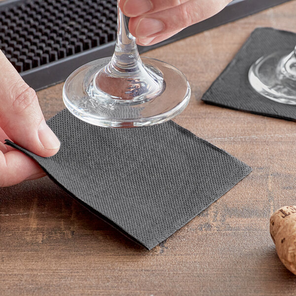 A hand holding a Hoffmaster black beverage napkin over a wine glass on a table.