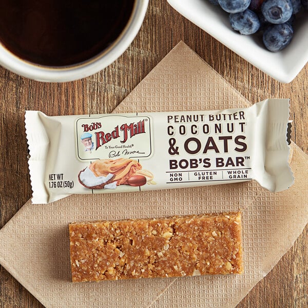 A brown Bob's Red Mill Peanut Butter Coconut & Oats bar on a table with a cup of coffee.
