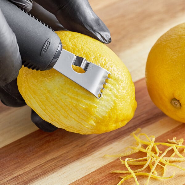 A person in black gloves using an OXO stainless steel citrus zester and channel knife to peel a lemon.