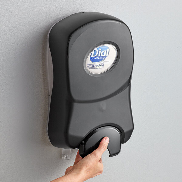 A hand pressing a Dial universal manual foaming hand soap dispenser on a wall.