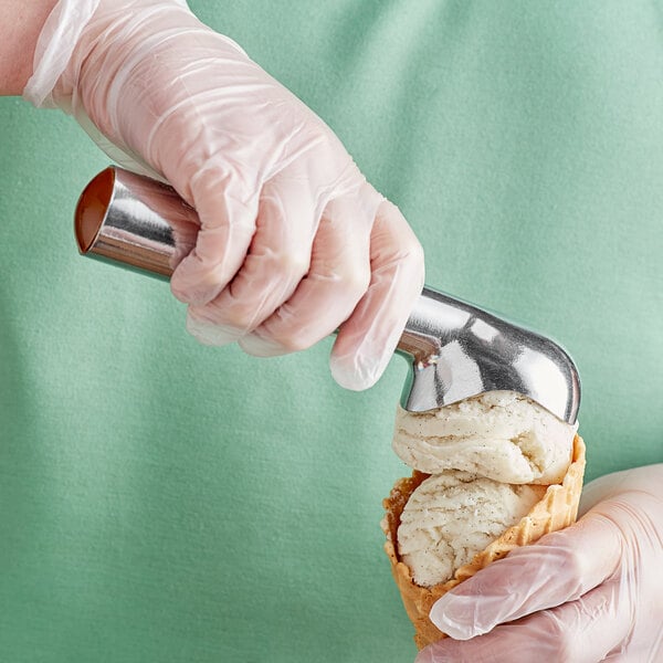 A person in gloves using a Choice aluminum gold ice cream scoop to fill a waffle cone.