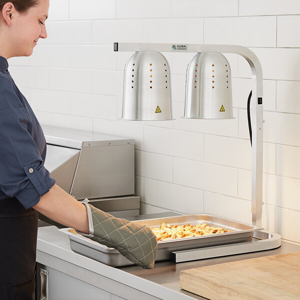A woman in a blue shirt and apron using a Global Solutions countertop heat lamp to keep food warm on a tray.