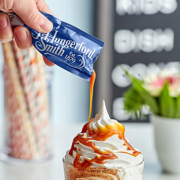 A hand pouring a J. Hungerford Smith Caramel packet into a glass of ice cream.