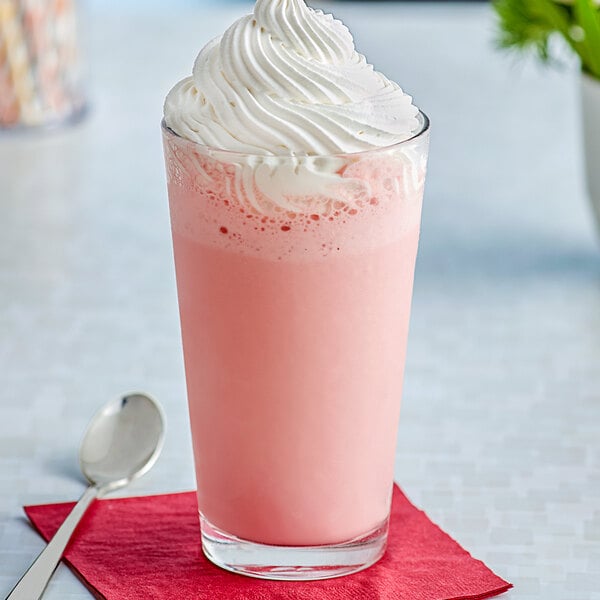 A glass of pink milkshake with whipped cream on top and a spoon on a napkin.