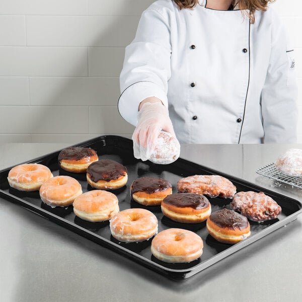 A chef in a black uniform places chocolate covered doughnuts on a Carlisle market tray.