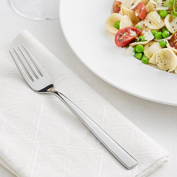 An Acopa stainless steel dinner fork on a napkin next to a plate of pasta with vegetables and cheese.