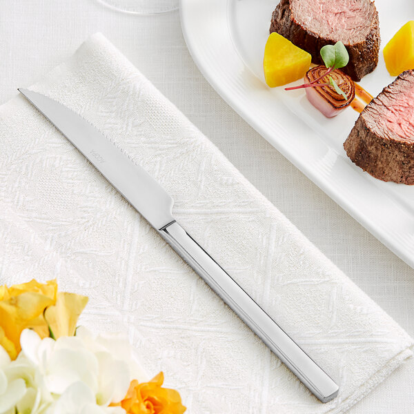 An Acopa stainless steel steak knife on a napkin next to a plate of meat and vegetables.