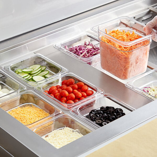 A gloved hand using a Vigor 1/6 size clear food pan to serve shredded carrots over a counter with bowls of tomatoes and vegetables.