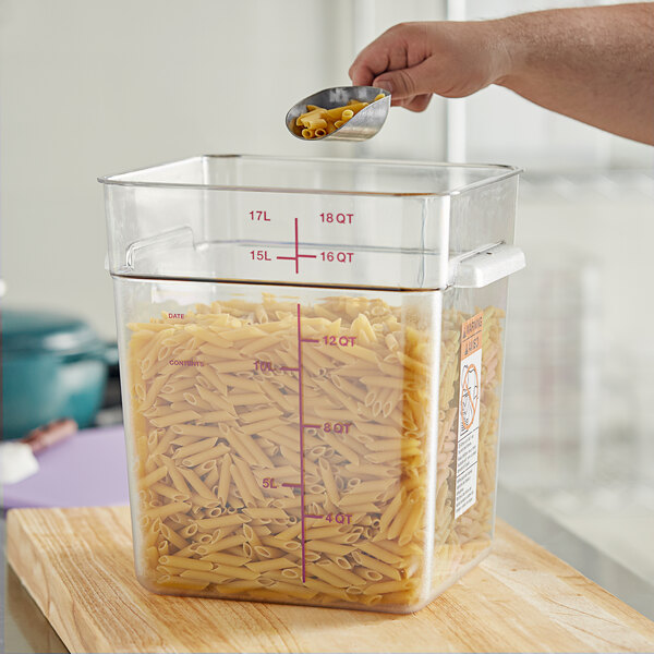 A hand using a measuring spoon to pour pasta into a large clear plastic container.