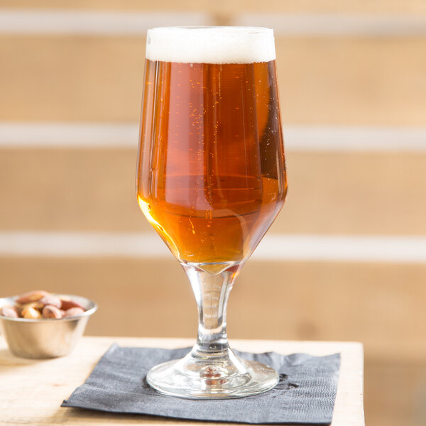 A close up of a Libbey stemmed pilsner glass of beer on a table with a bowl of nuts.