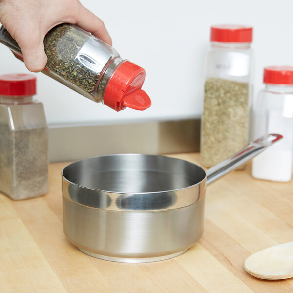 A hand holding a bottle of seasoning pouring into a Vollrath Centurion saute pan with a silver bowl of spices on a wood surface.