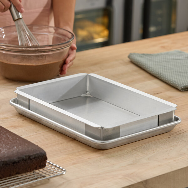 A rectangular metal baking pan with straight sides is placed on top of a matching metal tray on a wooden countertop, with a person whisking chocolate batter in a glass bowl nearby.