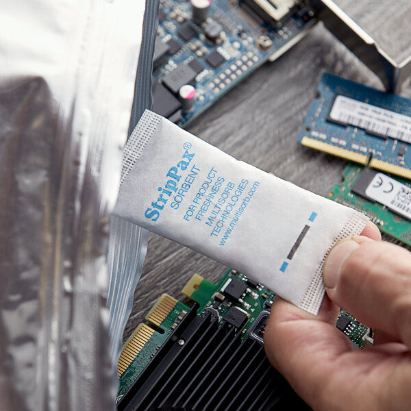 A hand holding a Multisorb StripPax desiccant packet over a circuit board.