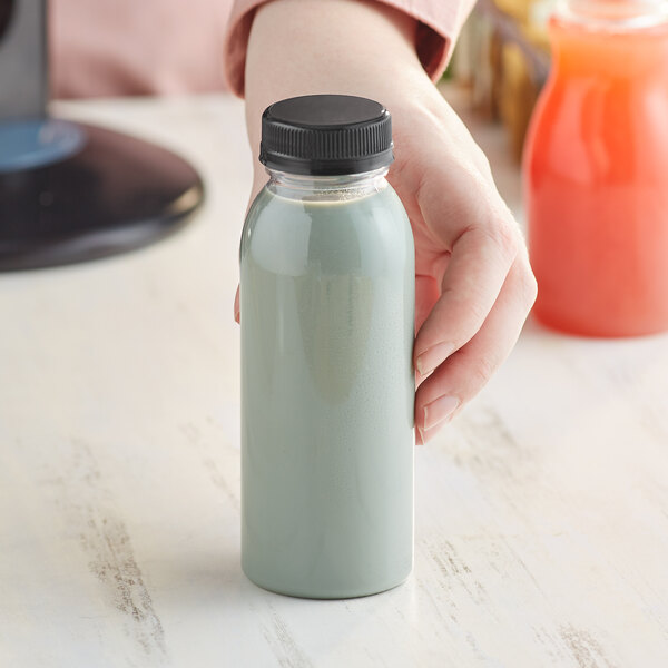 A hand holding an 8 oz. round clear PET juice bottle with green liquid and a black lid.