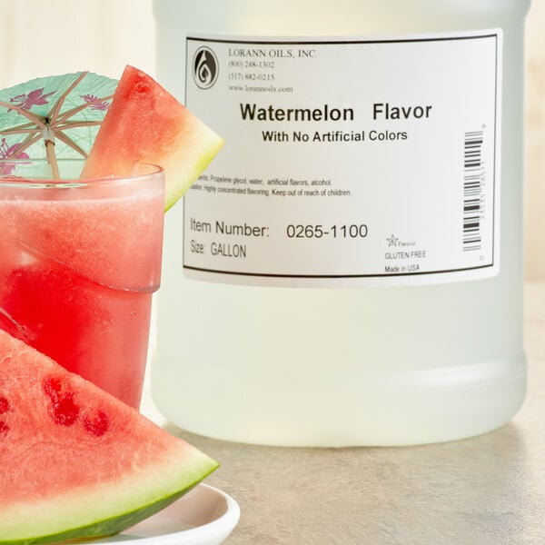 A glass of watermelon juice with a slice of watermelon on a plate next to a bottle of LorAnn Oils Watermelon Flavor.