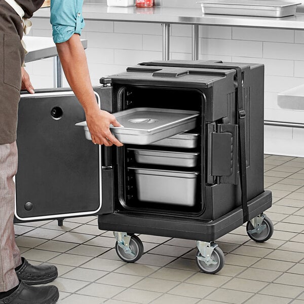 A man in a chef's uniform using a black Choice dolly to hold a metal container of food.