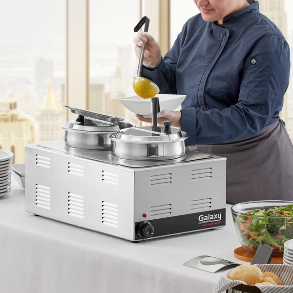 A woman in a chef's uniform using a Galaxy electric countertop food warmer to prepare food.