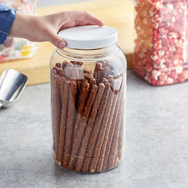 A hand holding a 1 gallon round PET plastic jar with a white lid filled with pretzels.