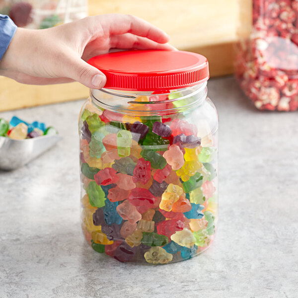 A hand holding a 64 oz. round PET plastic jar filled with gummy bears with a red lid.