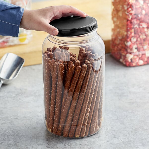 A person holding a 1 gallon plastic jar with a black lid filled with pretzels.