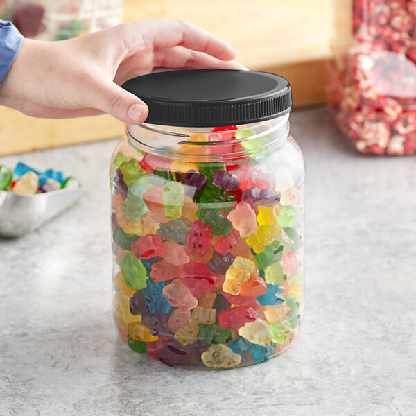 A hand holding a 64 oz. round PET plastic jar of gummy bears with a black lid.
