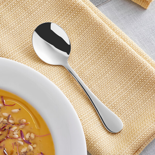 An Acopa Vittoria stainless steel bouillon spoon on a plate with a bowl of soup.