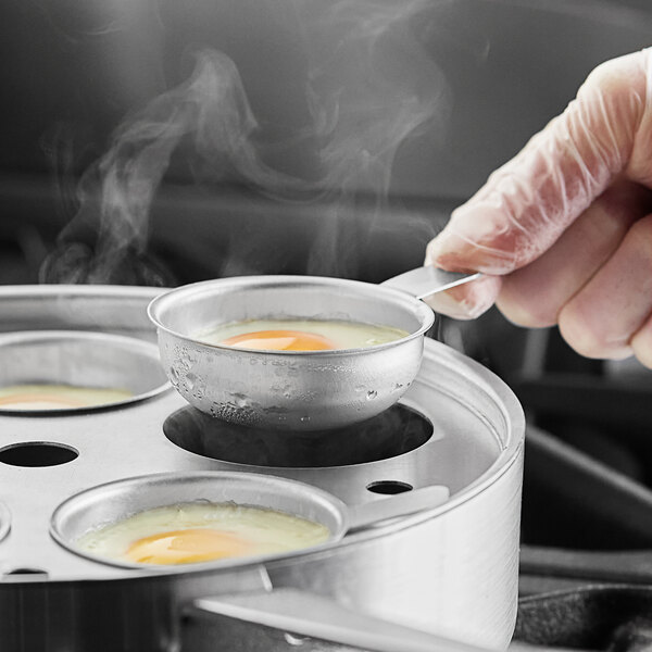 A person putting a Choice Natural Finish Aluminum Egg Poacher Cup into a pan with eggs.