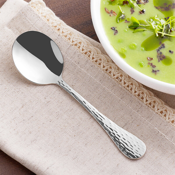 An Acopa Inspira stainless steel bouillon spoon on a napkin next to a bowl of green soup.