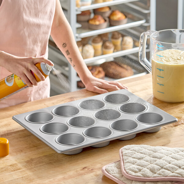 A 12-cup glazed aluminized steel jumbo muffin or cupcake pan being sprayed with nonstick spray on a kitchen counter.