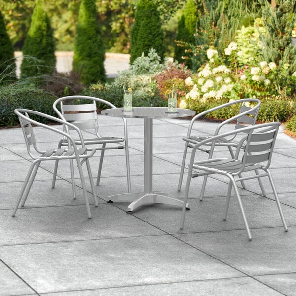 A Lancaster Table & Seating chrome round outdoor table with four silver arm chairs on a concrete patio.