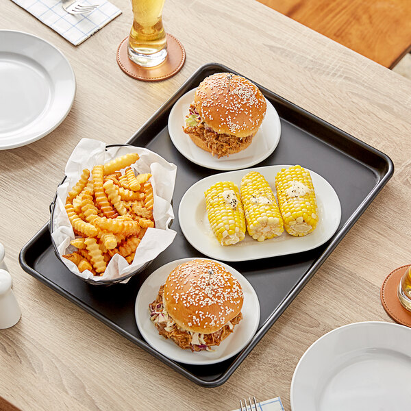 A Baker's Mark black non-stick bun tray on a wood table with a burger, fries, and corn.