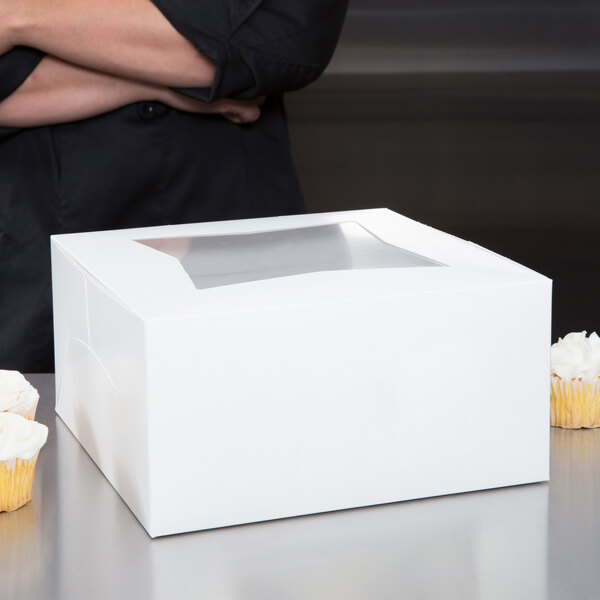 A person standing next to a white box with a clear window next to cupcakes.