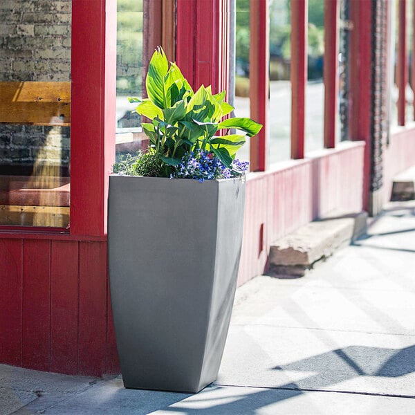 A Mayne graphite grey planter with flowers and leaves on a sidewalk.