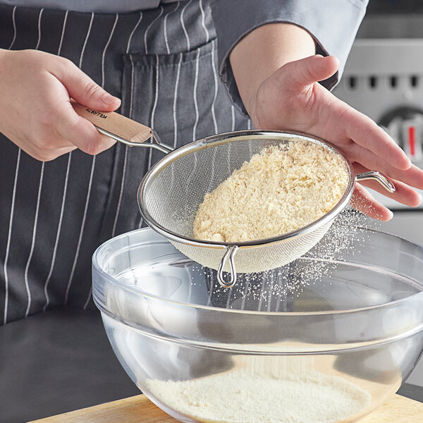 A person using a Vollrath single mesh strainer with a wood handle to sift flour into a bowl.