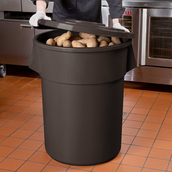 A man in a chef's uniform holding a large brown round ingredient storage bin with a lid full of potatoes.