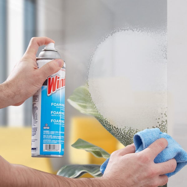 A person's hand spraying a window with a blue and white SC Johnson Windex aerosol container.