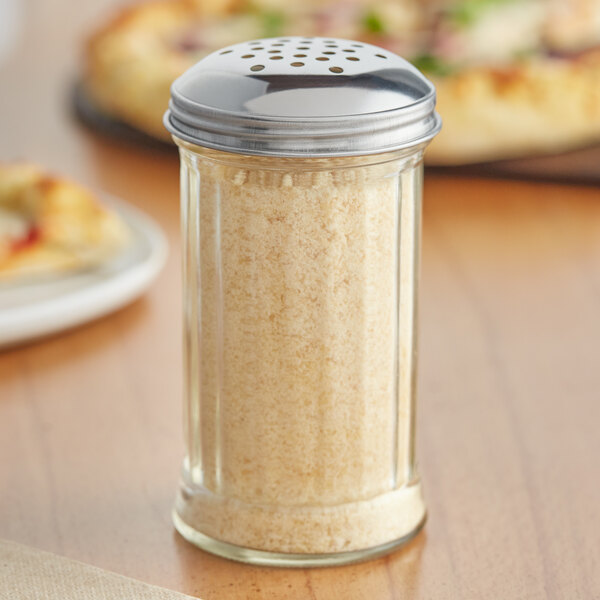 A Tablecraft glass cheese shaker with a silver lid on a table next to a pizza.